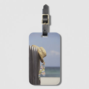 Strohhut, der am Zaun am Strand hängt Gepäckanhänger